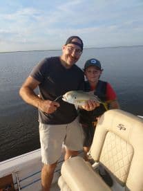 A proud angler and a young boy displaying a caught fish on a boat in a calm fishing environment.