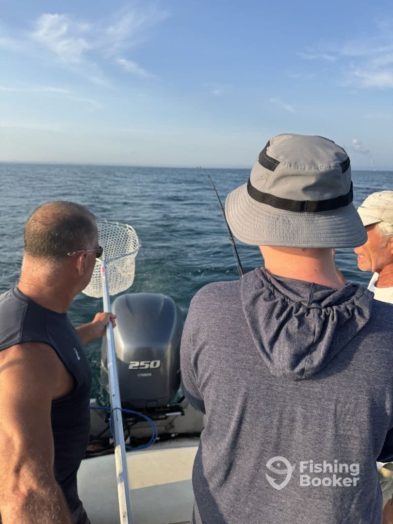 Three anglers engaged in fishing on a boat, with one preparing to net a catch in open water.