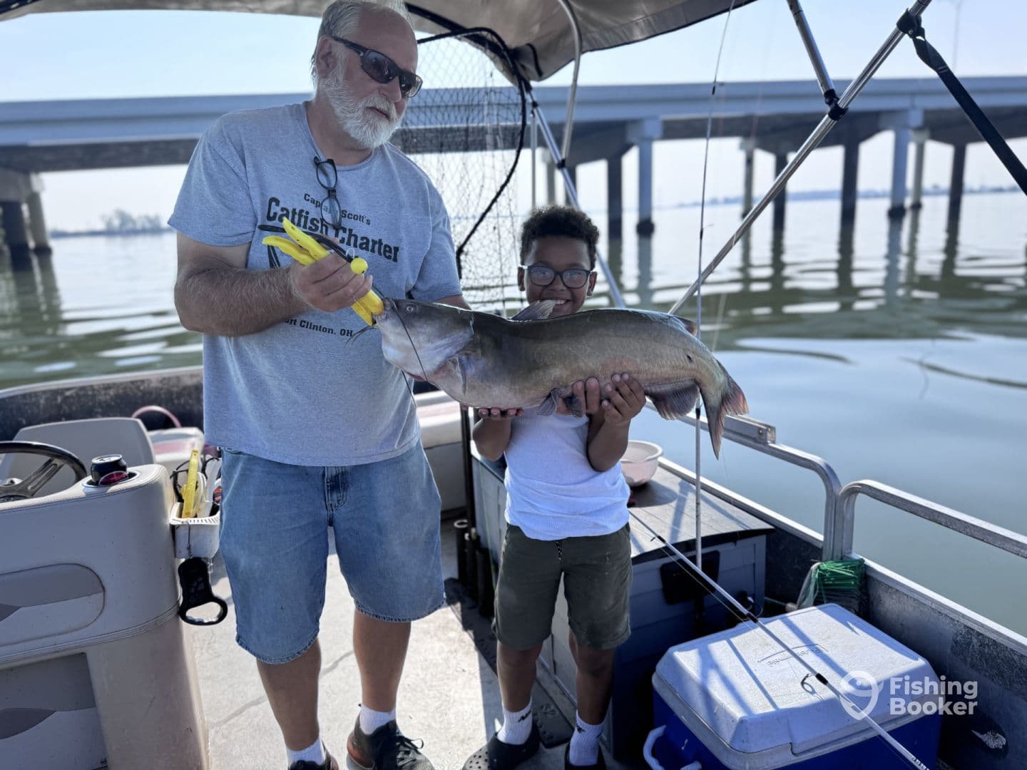 A young boy proudly holds a large Catfish while an adult assists him on a fishing charter in a calm water setting.