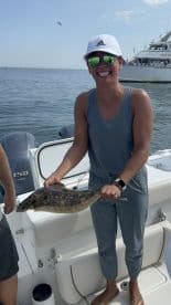 An angler proudly displaying a Flounder while on a fishing trip, with a boat in the background.
