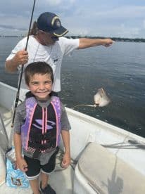 A young boy in a life jacket smiles broadly while fishing on a boat, with an adult angler in the background holding up a caught fish.