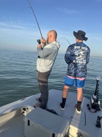 Two anglers engaged in fishing on a boat, with one reeling in a catch in calm waters under a clear sky.