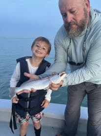 A young boy proudly poses with a Speckled Trout, assisted by an adult, while fishing on a boat.