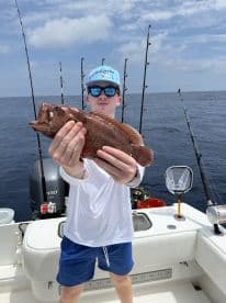 A young angler proudly displaying a Red Grouper while fishing offshore, surrounded by fishing rods on a charter boat.