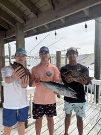 Three young anglers proudly displaying their catch, including a large King Mackerel and Snapper, at a marina after a successful fishing trip.
