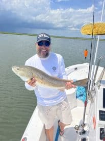 An angler proudly displaying a large Redfish while fishing on a boat in a scenic coastal environment.
