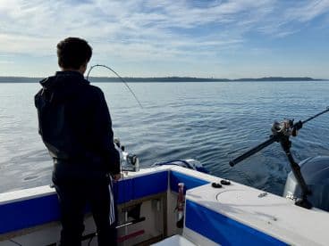 A young angler is actively reeling in a fish while fishing from a boat in calm waters, with a scenic backdrop of distant land.