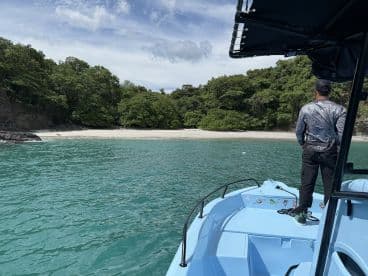 A lone angler stands on the bow of a light blue boat, gazing towards a secluded beach surrounded by lush greenery, showcasing a tranquil fishing environment.