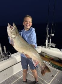 A young angler proudly displaying a large Lake Trout aboard a fishing boat during a night trip.