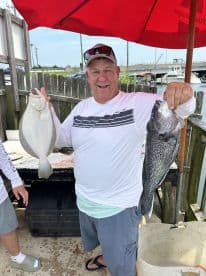 An enthusiastic angler proudly displaying a Flounder and a Black Sea Bass at a dockside cleaning station after a successful fishing trip.