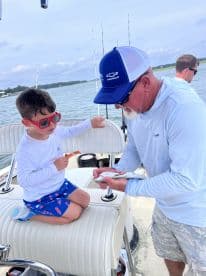 A young boy in red sunglasses watches intently as an adult shows him a freshly caught fish on a fishing boat, highlighting a memorable fishing experience.