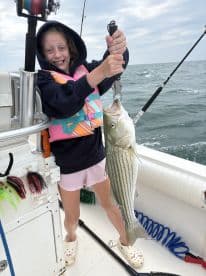 A young angler joyfully displaying a large striped bass while fishing on a boat, showcasing a fun day on the water.