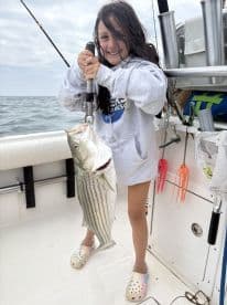 A young angler proudly displaying a large Striped Bass while fishing on a boat, showcasing a fun day on the water.