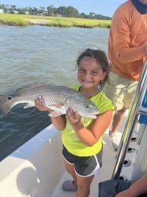 A young girl proudly displaying her catch, a Redfish, while fishing on a boat in a scenic coastal environment.