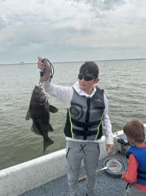 A young angler proudly displaying a large Black Drum while fishing on a boat, with another child observing nearby.