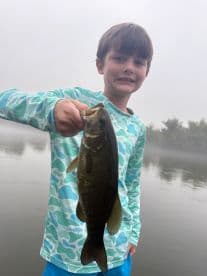 A young angler proudly displaying a caught Largemouth Bass while fishing on a misty lake.