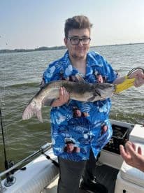 A young angler proudly displaying a large catfish while fishing on a sunny day, showcasing a vibrant fishing environment.