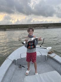 A young angler proudly displaying a Speckled Trout while fishing from a small boat, showcasing a beautiful sunset backdrop.
