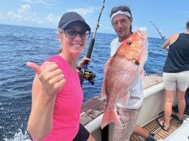 A woman gives a thumbs up while holding a fishing rod, alongside a man proudly displaying a large Red Snapper on a fishing charter in open waters.