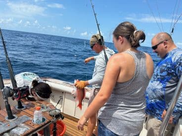 A group of anglers actively fishing on a boat, with one person reeling in a Red Snapper while others assist and observe in a sunny offshore setting.