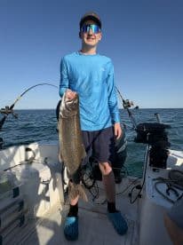 A young angler proudly displaying a large Lake Trout while fishing on a boat, showcasing a successful day on the water.