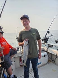 A young angler proudly displaying a large Salmon while fishing on a boat, with another angler visible in the background.