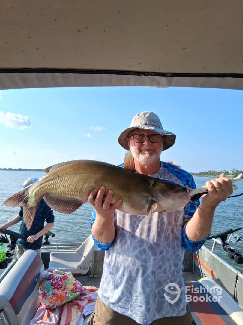 An angler proudly displaying a large catfish while fishing on a boat, with a scenic water backdrop.