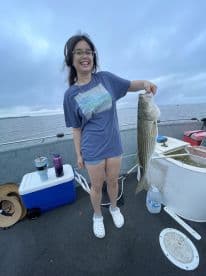 A happy angler holding a large Striped Bass while fishing on a boat, showcasing a successful catch during a serene outing.