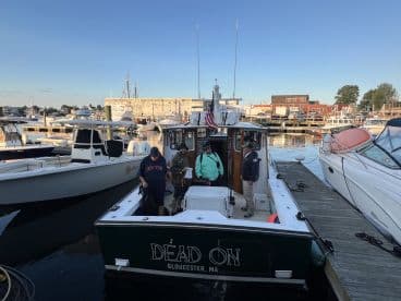 The 'Dead On' charter boat docked in Gloucester, MA, with crew members preparing for a fishing trip amidst a bustling marina.