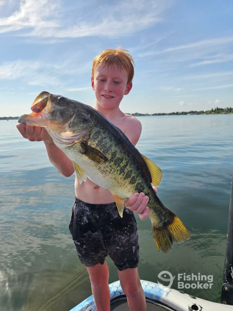 A young angler proudly displaying a large Largemouth Bass while fishing on a calm lake.