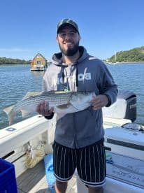 Angler proudly displaying a large Striped Bass while on a boat, with a scenic waterfront in the background.