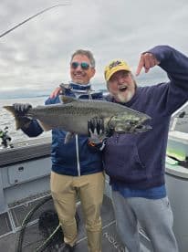 Two anglers proudly displaying a large Chinook Salmon on a fishing boat, showcasing a successful day on the water.