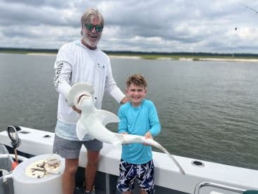 A young boy and an adult proudly display a Hammerhead Shark on a fishing boat, showcasing a successful catch during a family fishing trip.