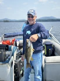 An adult angler proudly displaying a large Smallmouth Bass while fishing on a serene lake, surrounded by mountainous scenery.