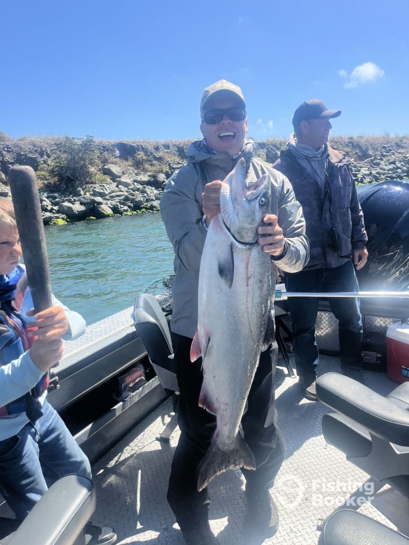 A proud angler displaying a large Salmon while fishing with family on a boat, surrounded by a scenic river landscape.