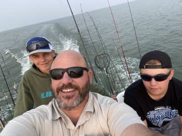 A father and two young anglers enjoying a day on the water, seated inside the boat with fishing rods in the background.