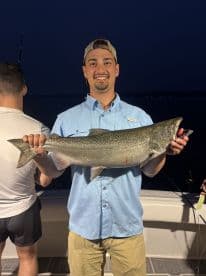 An angler proudly displaying a large Salmon on a fishing boat at night, showcasing a successful catch during a fishing trip.