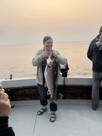 A smiling angler proudly displaying a large lake trout on a fishing boat during sunset, showcasing a successful fishing trip.
