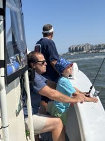 A young boy and an adult are actively fishing from a boat, enjoying a sunny day on the water, with a scenic backdrop of coastal buildings.