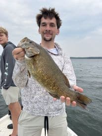 Angler proudly displaying a large Largemouth Bass while fishing on a lake, with another person in the background.