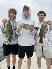 Three young anglers proudly displaying their catch of Smallmouth Bass while fishing on a boat.