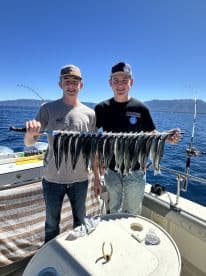 Two young anglers proudly displaying a catch of multiple fish on a sunny day while fishing on a boat, with scenic mountains in the background.