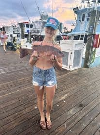 A young angler proudly displaying a Snapper caught during a fishing trip at the marina, with boats in the background.