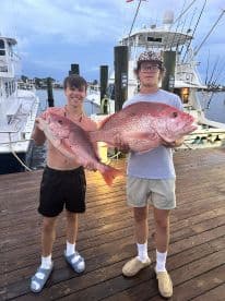 Two young anglers proudly displaying their large Red Snapper catch at the dock, showcasing a successful fishing trip.