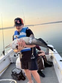 A young angler proudly holding a large catfish while fishing on a boat during sunset.