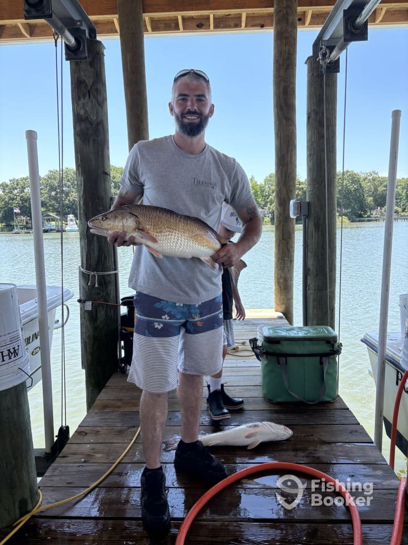 An angler proudly displaying a large Redfish on the dock, showcasing a successful day of fishing in a scenic waterfront setting.