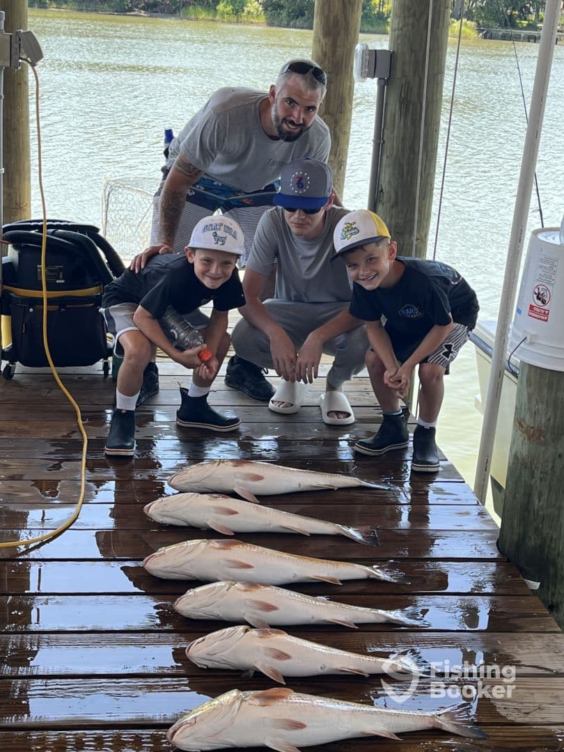 A group of young anglers and an adult proudly displaying their catch of several large fish on a dock, showcasing a successful fishing trip.