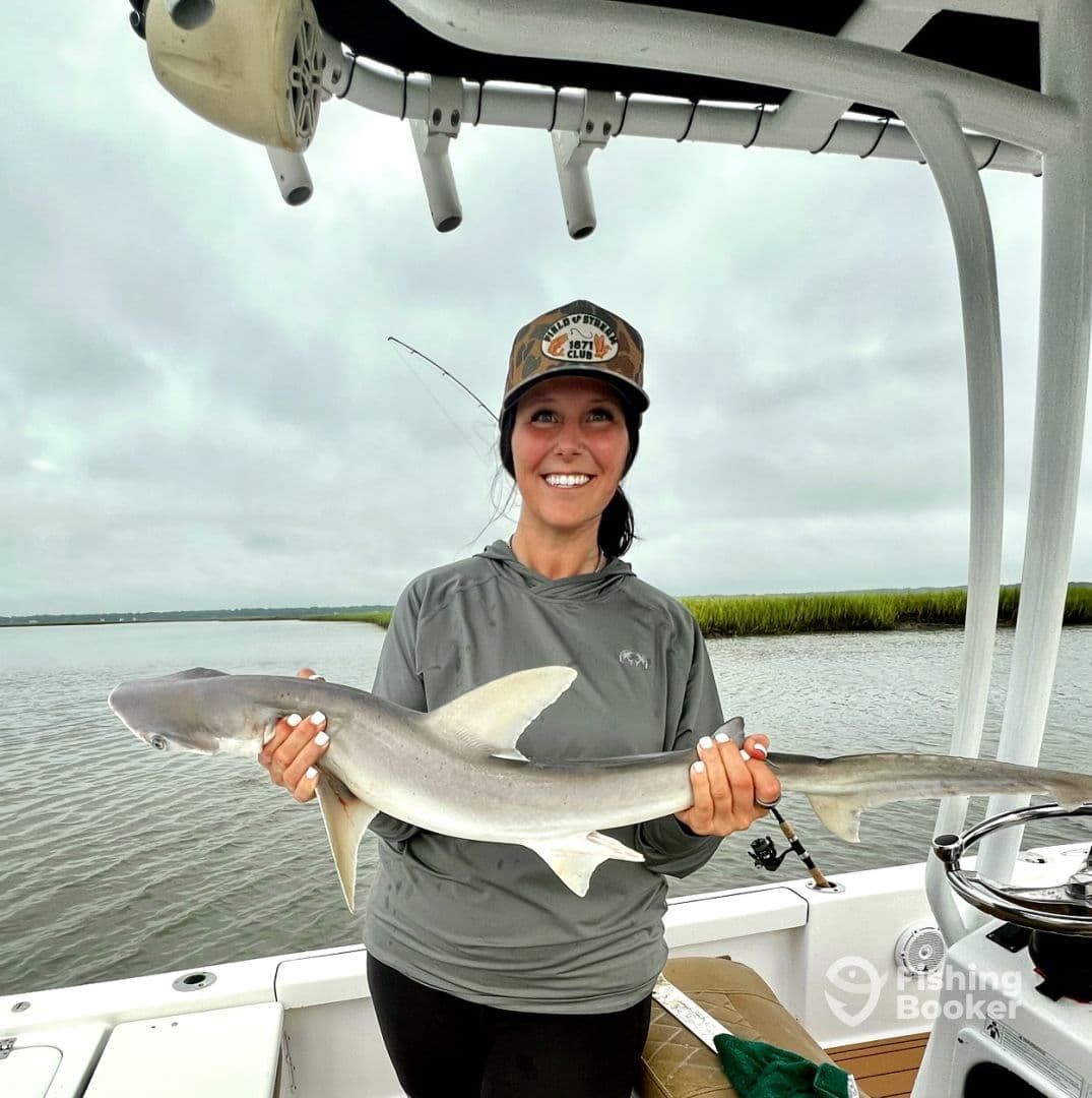 A smiling angler proudly displays a caught shark while on a fishing trip, showcasing the serene waters and lush greenery in the background.