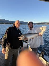 Two anglers proudly displaying a large Lake Trout while fishing on a scenic lake, surrounded by mountains.