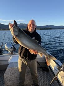 An angler proudly displaying a large Lake Trout while fishing on a scenic lake, surrounded by mountains.